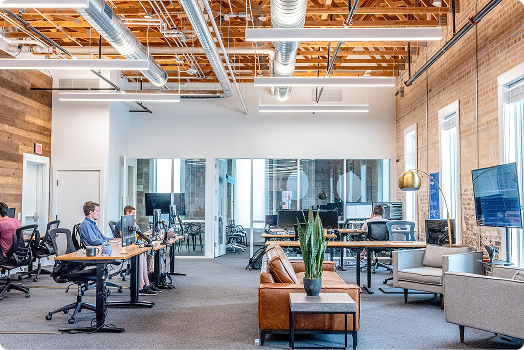 Modern open-plan office with exposed ceilings, desks with computers, several people working, glass-walled meeting rooms, and a lounge area with a sofa and plants.