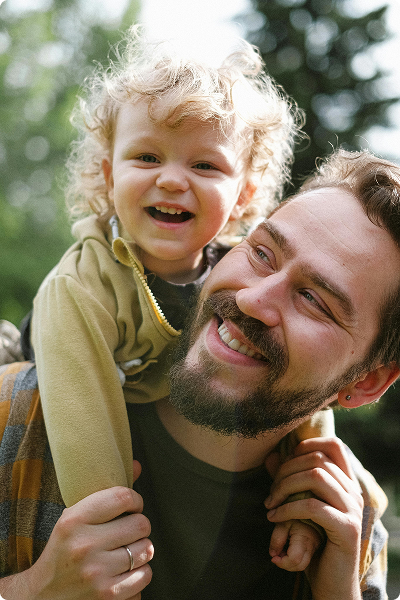 A smiling man carries a laughing toddler with curly hair on his shoulders outdoors, with trees blurred in the background.