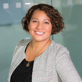 Woman with short curly hair wearing a striped blazer and black top, smiling, standing in front of a glass wall with square patterns.