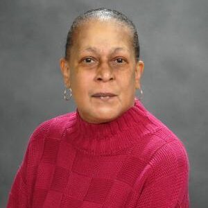 A woman with short hair, wearing hoop earrings and a textured magenta sweater, poses in front of a gray, studio-style background.