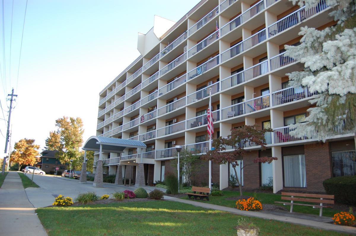 A multi-story apartment building with balconies, an entrance canopy, a sidewalk, a flagpole, benches, and landscaped greenery in front.