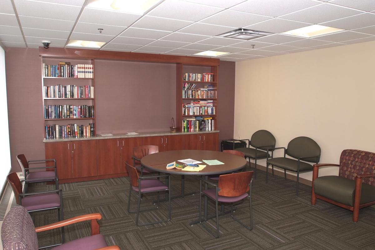 Small conference room with a round table, six chairs, bookshelves filled with books, and papers on the table. The room has beige walls and an overhead fluorescent light.