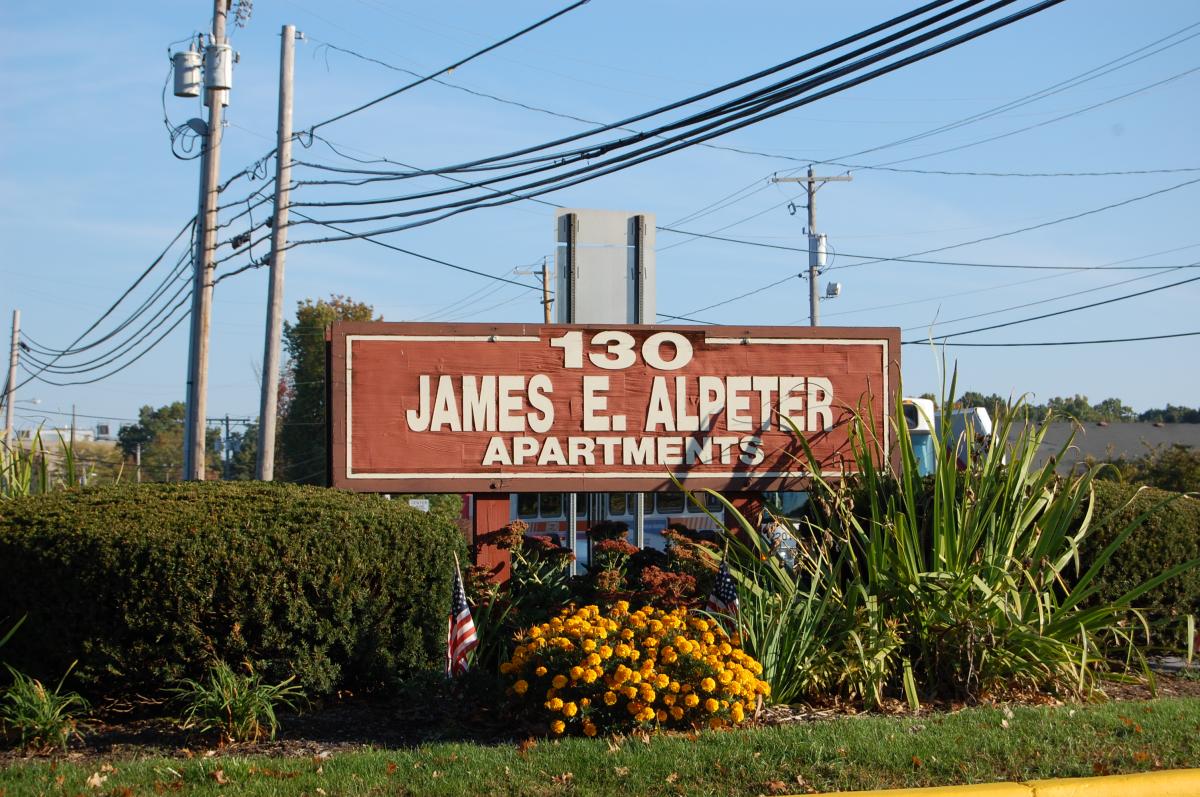 A brown sign reads "130 James E. Alpeter Apartments" surrounded by bushes, flowers, and utility poles in the background.
