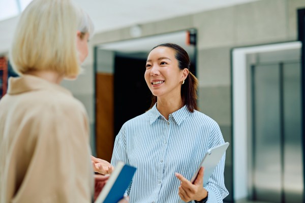 Two women stand indoors having a conversation; one holds a tablet, and both appear engaged in discussion. An elevator and a blurred background are visible behind them.