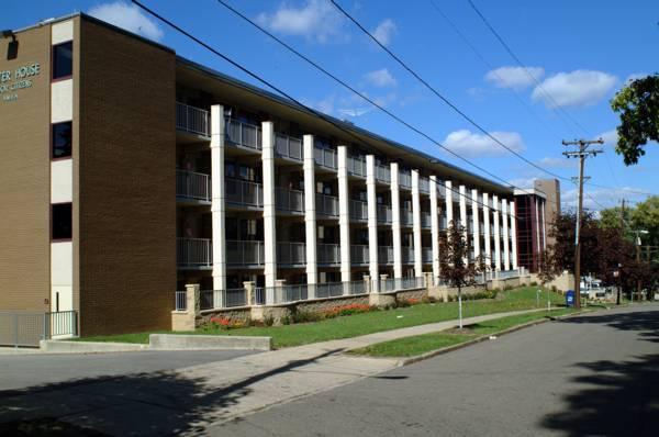 A four-story apartment building with balconies and a brick exterior, situated on a tree-lined street under a blue sky with scattered clouds.
