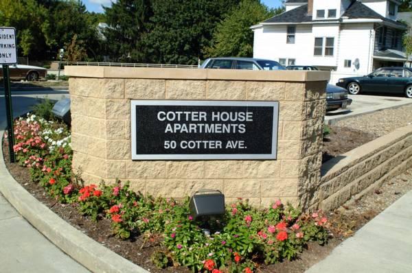 Outdoor sign reading "Cotter House Apartments, 50 Cotter Ave." is mounted on a brick wall with flowers planted in front.