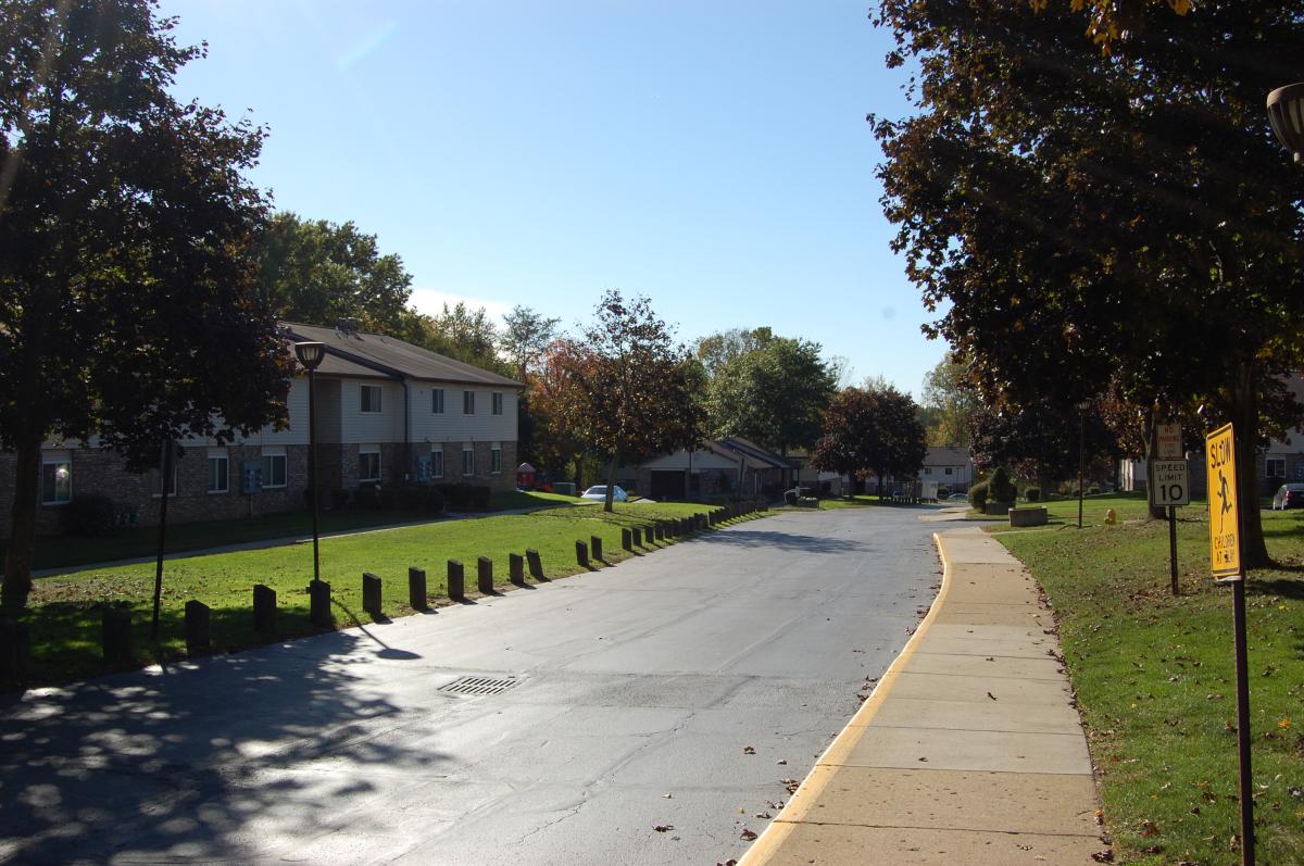 A quiet residential street with apartment buildings, trees, green lawns, a sidewalk, and a speed limit sign under a clear blue sky.