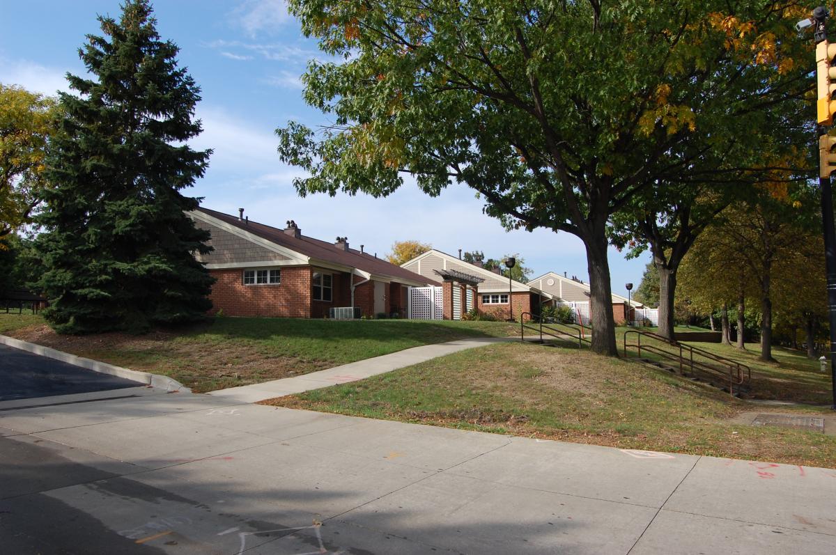 Single-story brick buildings with sloped roofs and chimneys sit beside a sidewalk and a sloped grassy area, bordered by trees and a paved street under a blue sky.