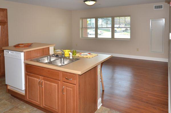 A kitchen with a double sink island, dishwasher, and wooden cabinets opens to an empty room with wood flooring and a large window.