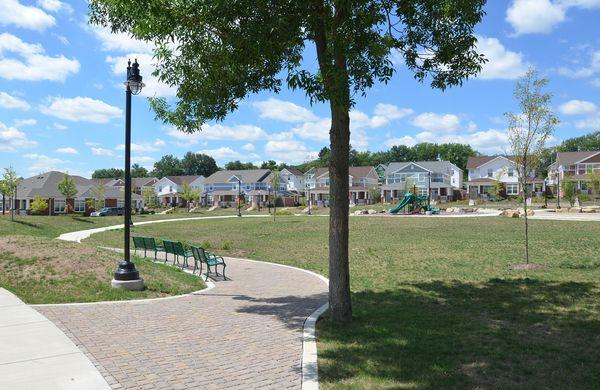 A paved path with benches curves through a grassy park, with a tree in the foreground and rows of suburban houses and a playground in the background under a blue sky with clouds.