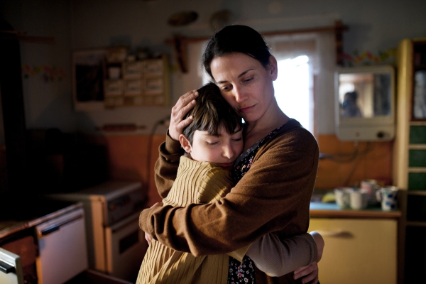 A woman hugs a child in a dimly lit kitchen, both with closed eyes, conveying comfort and affection.