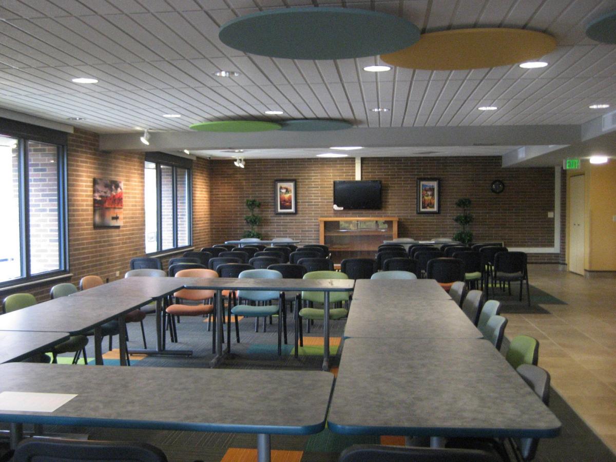 Modern meeting room with rectangular tables arranged in a U-shape, rows of chairs, large windows, wall-mounted TV, and ceiling panels.