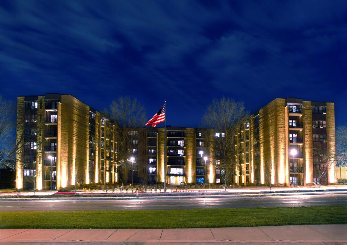 A large, illuminated apartment building with five floors stands at night, with an American flag in front and a road with light trails in the foreground.