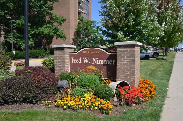 Brick sign reading "Fred W. Nimmer" surrounded by colorful flowers, with buildings, sidewalk, and trees in the background.