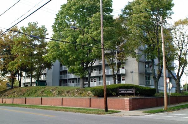 A multi-story apartment building with balconies, surrounded by trees and a brick wall, is seen from across the street on a clear day.