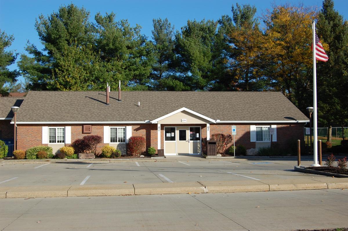 Single-story brick building with white trim, front entrance doors, empty parking spaces, landscaped shrubs, flagpole with American flag, and pine trees in the background under a clear sky.