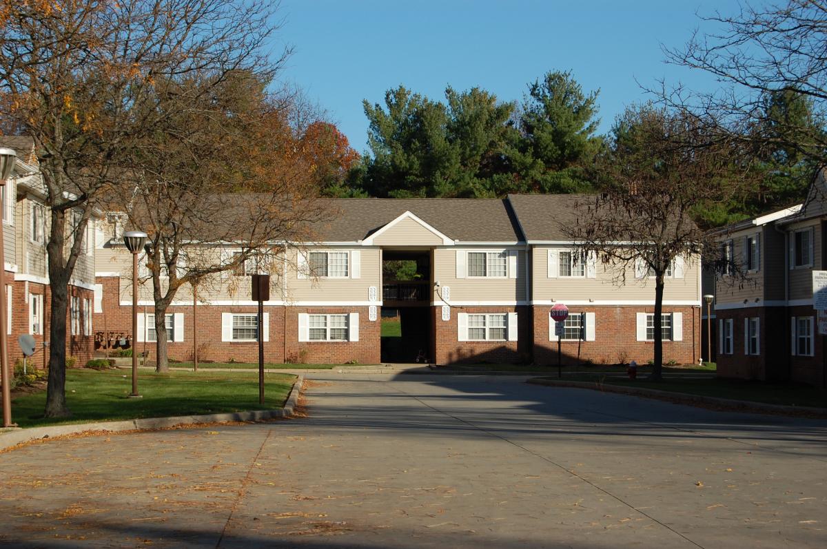 Two-story apartment buildings with brick and siding exteriors, facing a paved courtyard with some trees and green grass, under a clear blue sky.