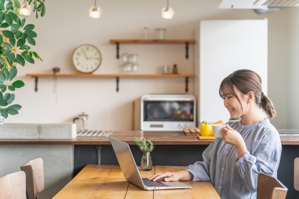 A woman sits at a kitchen table, holding a mug and using a laptop. The kitchen has shelves, a clock, and appliances visible in the background.