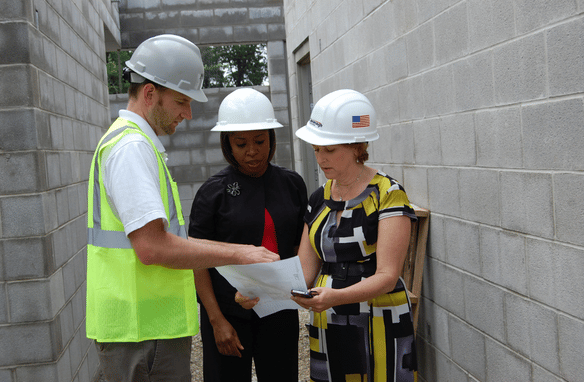 A plumber wearing safety goggles and a cap inspects pipes under a kitchen sink, writing notes on a clipboard.