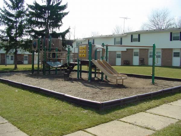 Playground with slides, a climbing structure, and swings, surrounded by grass and bordered by wooden timbers, situated near residential buildings.