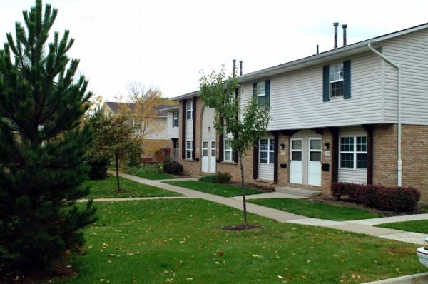 Two-story apartment buildings with brick and siding exteriors, small trees, shrubs, and sidewalks in a grassy courtyard setting.