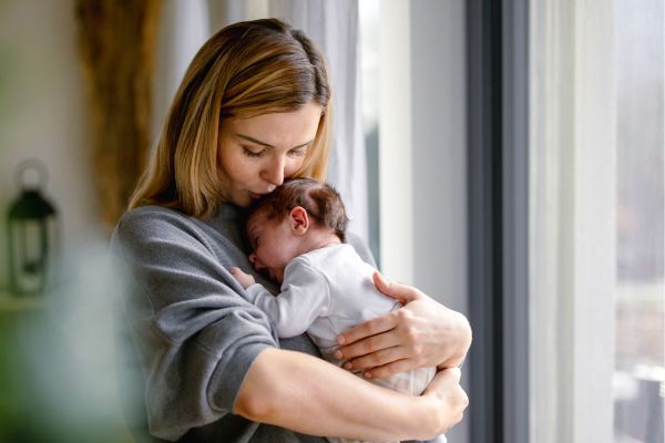 A woman holds and comforts a sleeping baby by a window during the daytime.