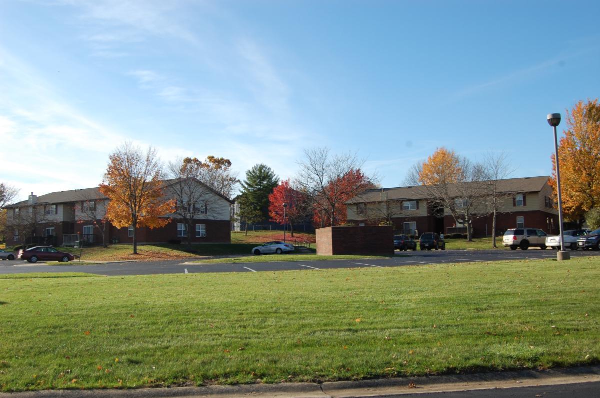 Apartment buildings with autumn trees and parked cars in front, seen across an empty parking lot and grassy area under a clear sky.