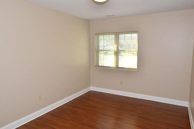 Empty room with beige walls, a window with blinds, white trim, and wood flooring. Ceiling light fixture is visible. Natural light enters through the window.