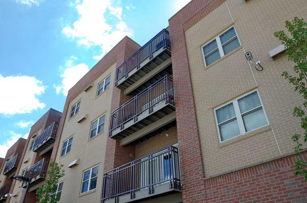Four-story modern apartment building with balconies, surrounded by green lawns, young trees, and a paved walkway under a blue sky with scattered clouds.