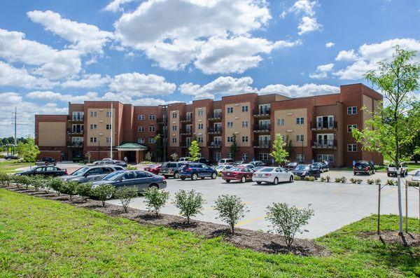 A four-story apartment building with a brick facade and balconies is shown behind a parking lot filled with cars under a partly cloudy sky.