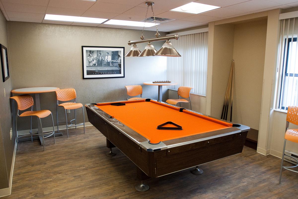Small game room with an orange pool table, orange chairs, two tall tables, a wall-mounted cue rack, wood floor, and a black-and-white photo on the wall.