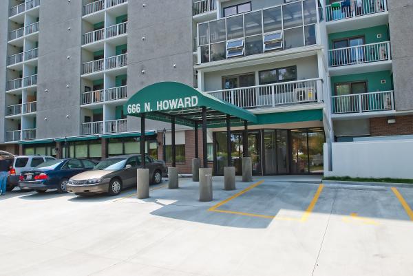 Entrance to a multi-story apartment building with a green awning labeled "666 N. Howard" over the driveway and parked cars nearby.