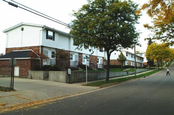 Row of two-story apartment buildings with brick and white siding, trees lining the sidewalk, and a person walking on the street in a residential neighborhood.