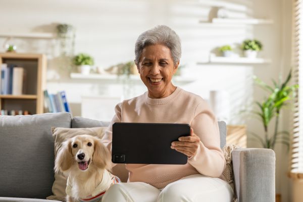 Older woman sitting on a couch, smiling while using a tablet, with a small dog beside her in a bright, modern living room.