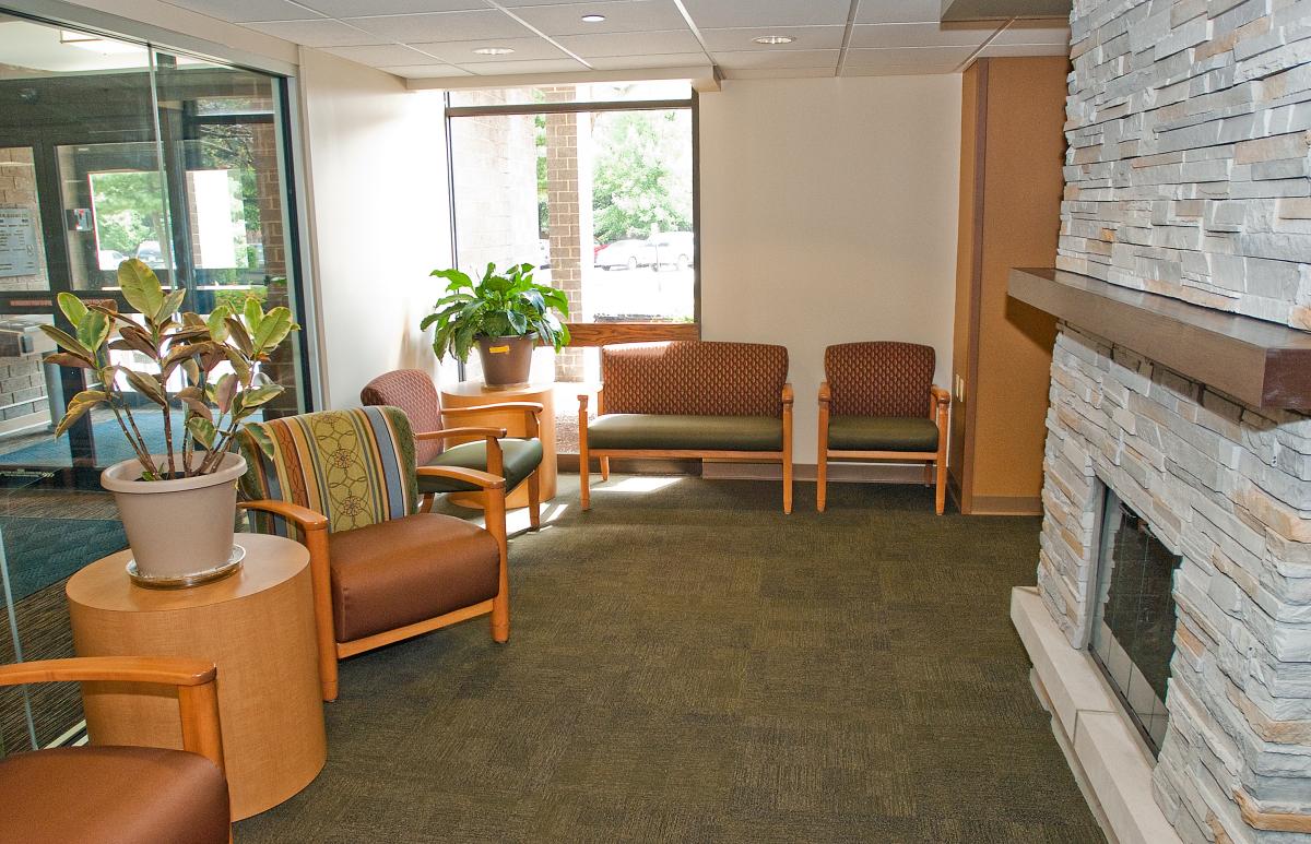 A waiting room with several chairs, a round table with a potted plant, large windows, and a stone fireplace on the right wall.