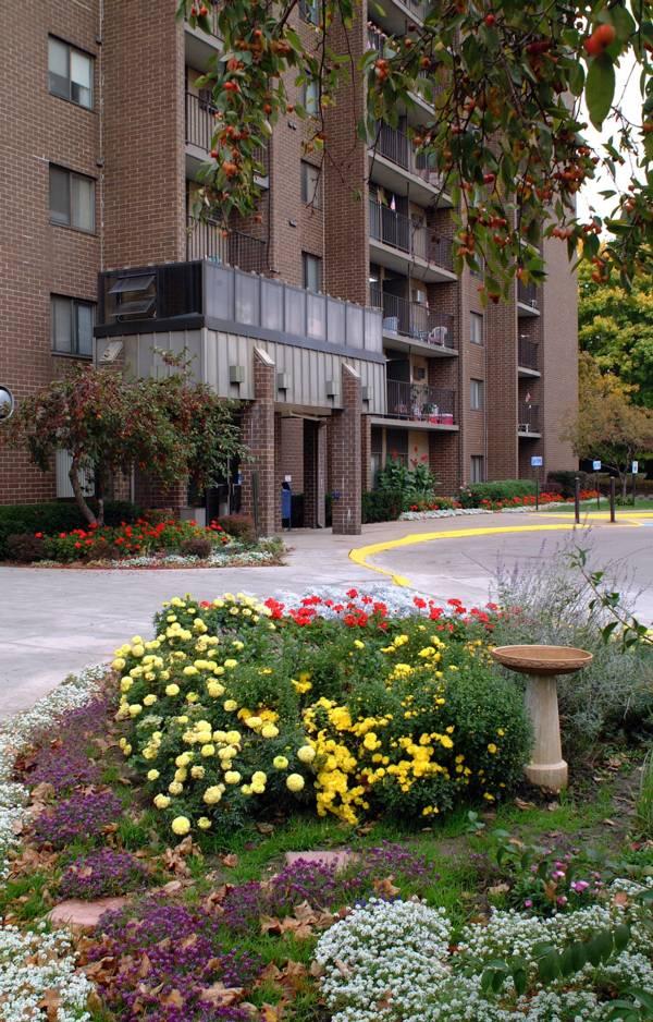 A brick apartment building with balconies, a landscaped garden with colorful flowers, and a curved driveway in front.