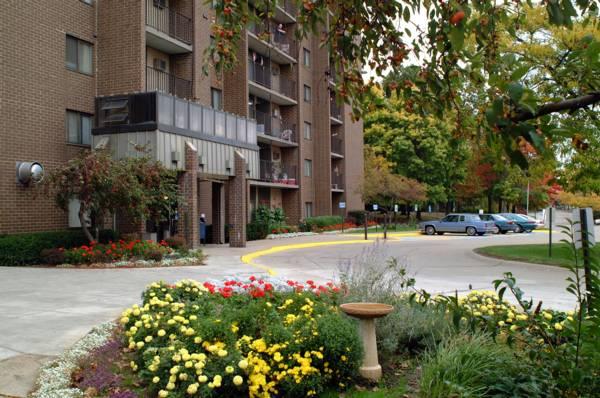 Brick apartment building with balconies, landscaped garden with yellow and red flowers, birdbath, and a curved driveway with parked cars in front. Trees with green foliage in the background.