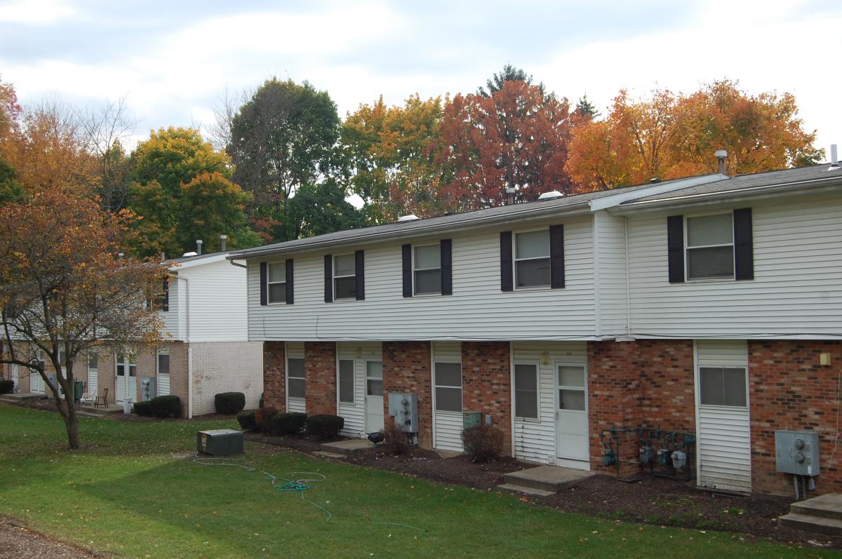 Two-story apartment buildings with brick and white siding exteriors, surrounded by grass and trees with autumn foliage.