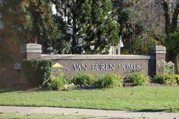 Brick sign with white letters reads "Van Buren Homes," surrounded by green shrubs and grass, with trees and a sidewalk in the background.