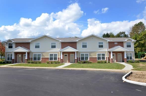 A row of two-story suburban townhouses with brick and beige siding, each with a front door, porch, and small lawn, under a partly cloudy sky.